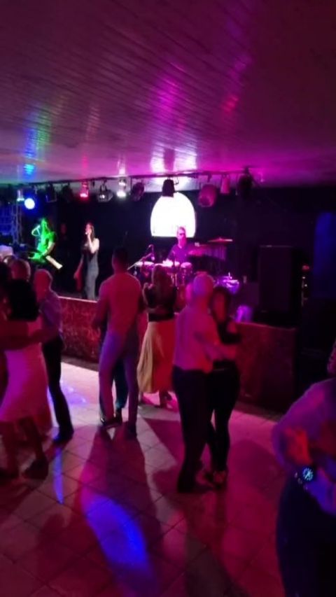 Pista de dança cheia, casais dançando sob luz rosa, com a banda tocando no palco ao fundo